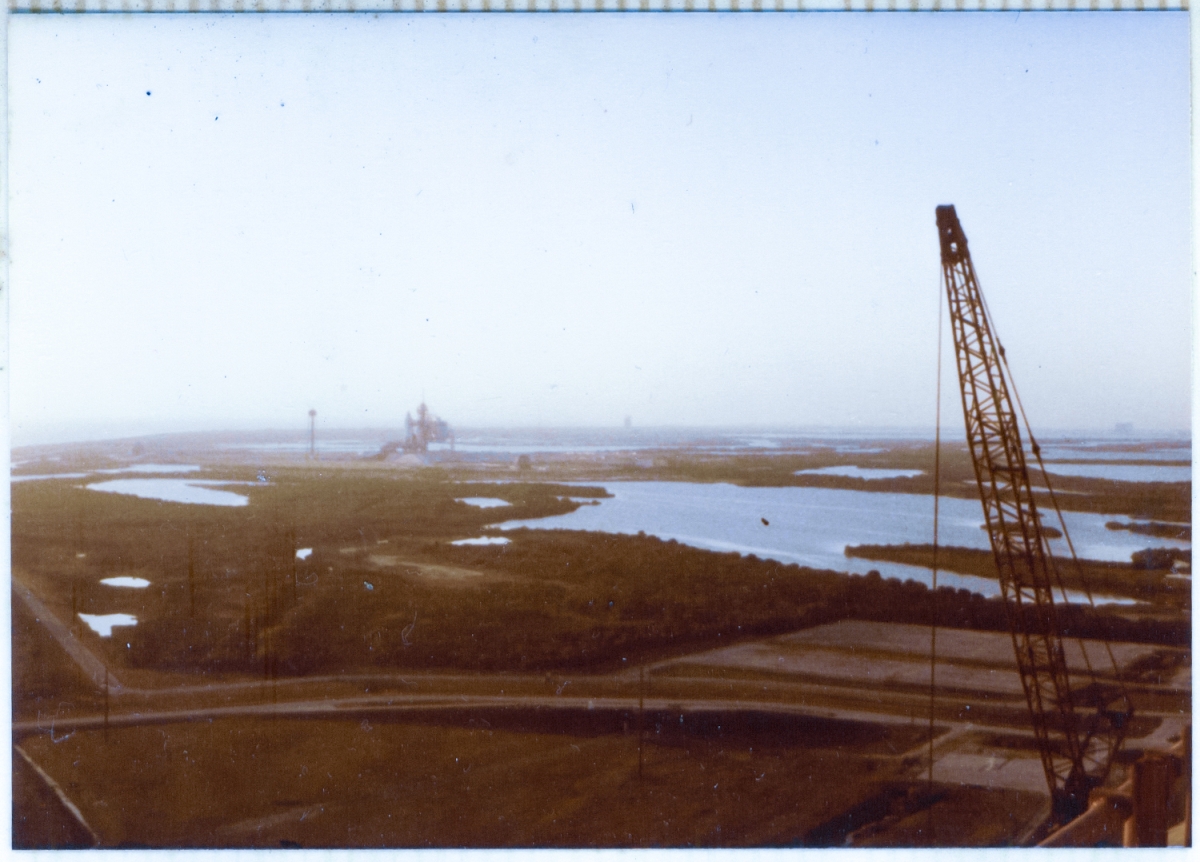 In the distance, across the wetlands of the Merritt Island Wildlife Refuge, Space Shuttle Columbia sits on top of Launch Complex 39-A, prior to taking flight on its second mission, STS-2. Your view is from near the top of the Fixed Service Structure at Pad-B, and the jib of the crane boom used to erect the steel elements of Pad-B can be seen on the right side of the image, along with a small portion of the handrail of the FSS at Pad-B, visible in the bottom right corner of the frame.